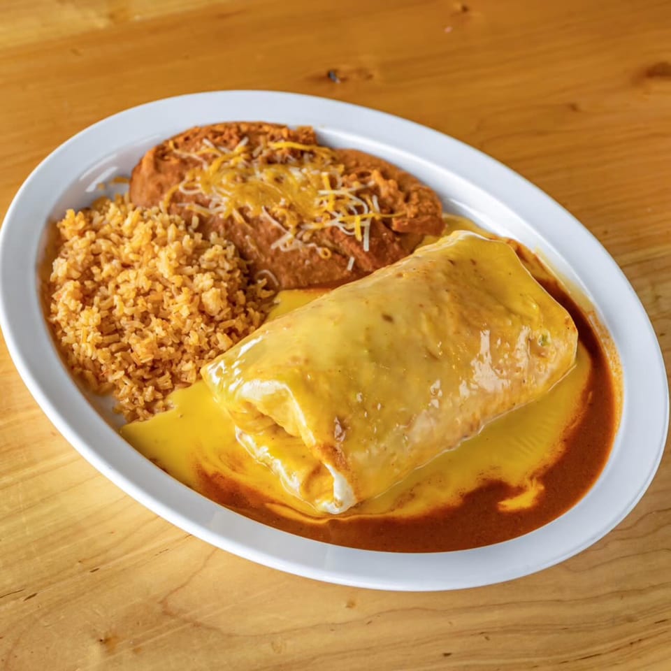 A Tacos El Cuñado Cascade burrito covered in cheese sauce, with rice and refried beans.